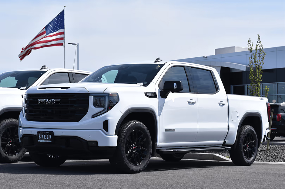 White GMC pickup truck with black wheels parked at dealership, American flag waving in background on clear day