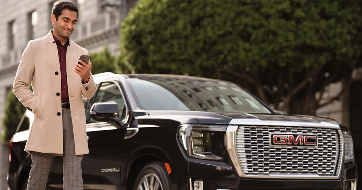 Man standing beside a black GMC Yukon Denali SUV using his smartphone with remote features, outside city buildings near Speck Buick GMC of Tri-Cities in Kennewick, WA.