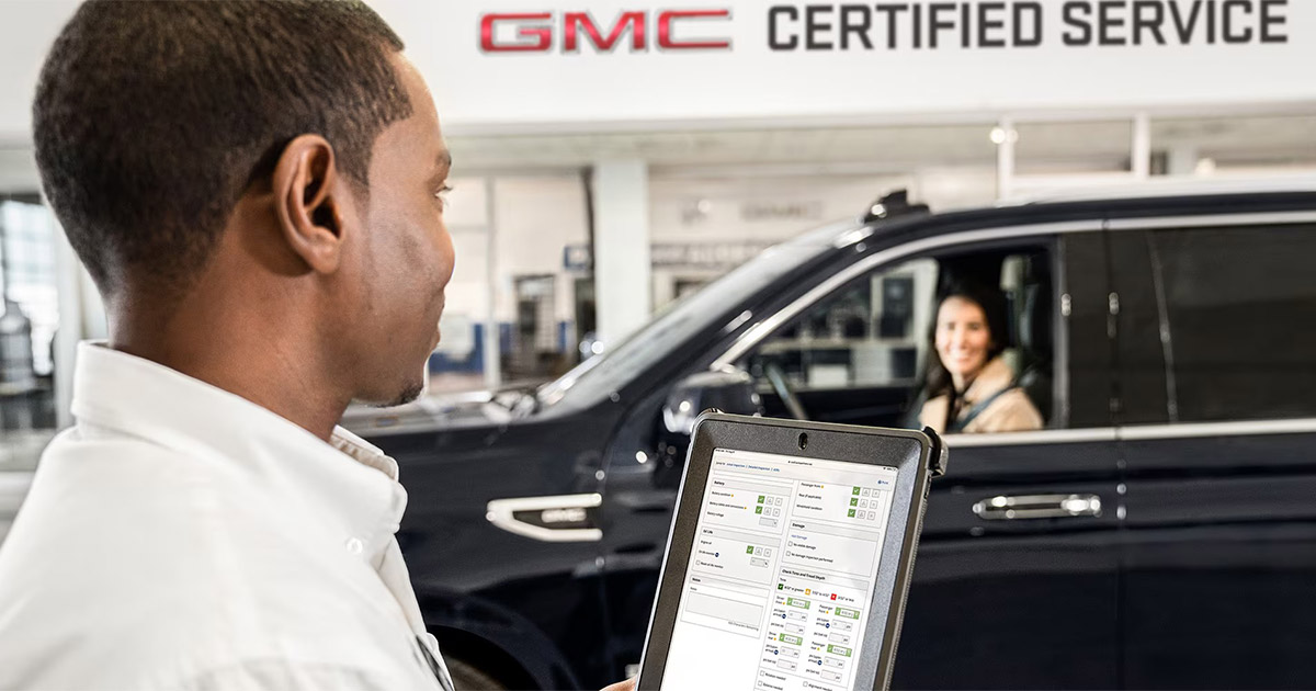 GMC certified service technician uses tablet to diagnose vehicle while customer sits in black SUV at dealership