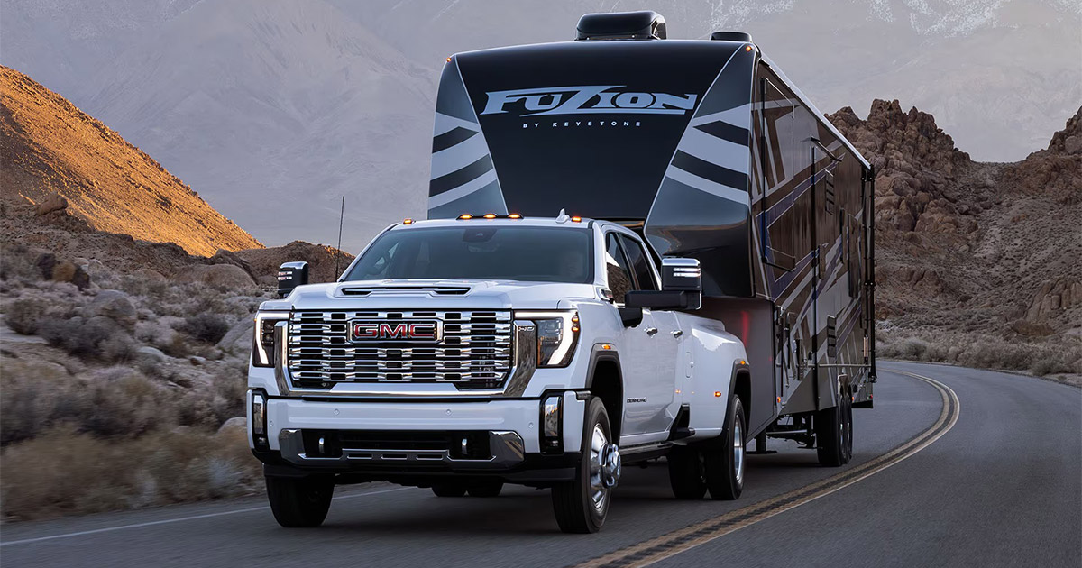 2025 GMC Sierra HD Denali in white towing a large fifth-wheel RV through a scenic mountain road, showcasing powerful towing capability, available at Speck Buick GMC of Tri-Cities in Kennewick, WA.