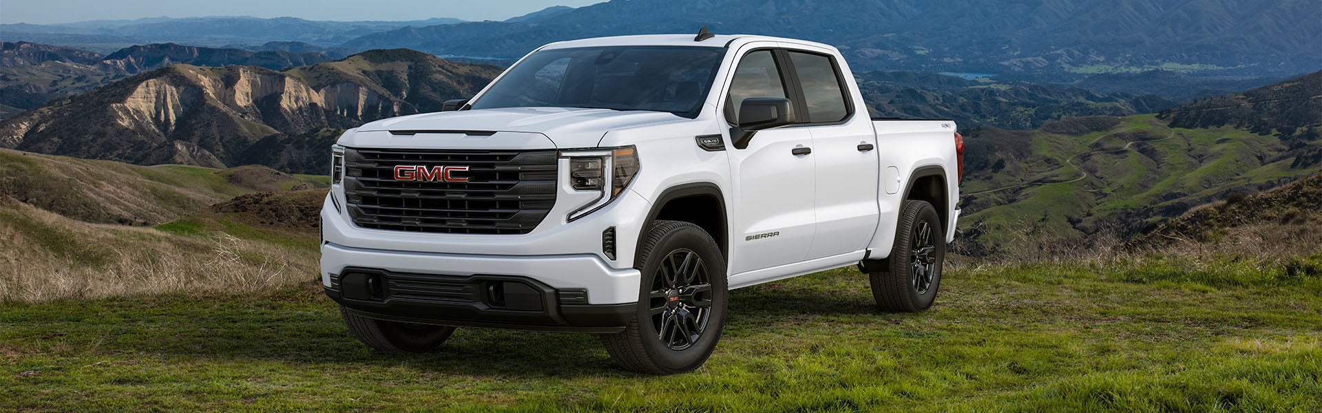 A white **GMC Sierra** pickup truck with black wheels parked in a grassy, hilly landscape.