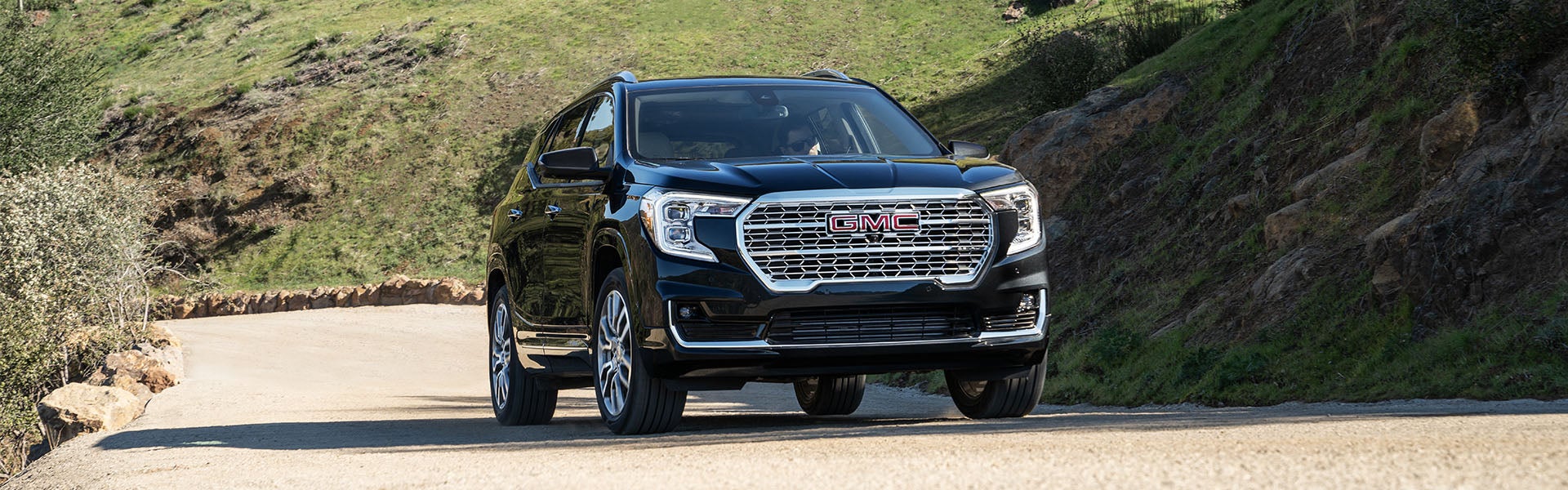 A black GMC Terrain SUV with a large chrome grille driving on a winding dirt road between grassy hills